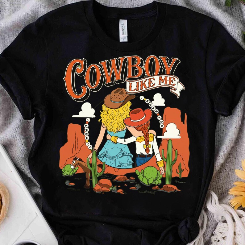 Puede incluir: Una camiseta negra de manga corta con un estampado gr&aacute;fico de dos figuras con sombreros de vaquero en un paisaje des&eacute;rtico con cactus y formaciones rocosas. El texto "COWBOY LIKE ME" aparece arriba, con "I KNEW YOU" en un borde circular.