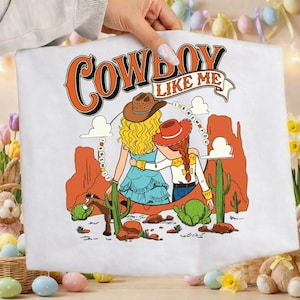 Puede incluir: Una camiseta blanca con una ilustraci&oacute;n gr&aacute;fica de dos figuras con sombreros de vaquero en un paisaje des&eacute;rtico con formaciones rocosas rojas, cactus y un caballo. El texto COWBOY LIKE ME e I KNEW IT I KNEW YOU est&aacute; impreso en naranja y negro.