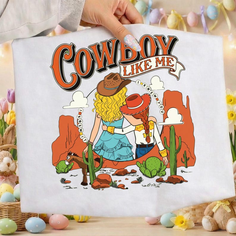 Puede incluir: Una camiseta blanca con una ilustraci&oacute;n gr&aacute;fica de dos figuras con sombreros de vaquero en un paisaje des&eacute;rtico con formaciones rocosas rojas, cactus y un caballo. El texto COWBOY LIKE ME e I KNEW IT I KNEW YOU est&aacute; impreso en naranja y negro.