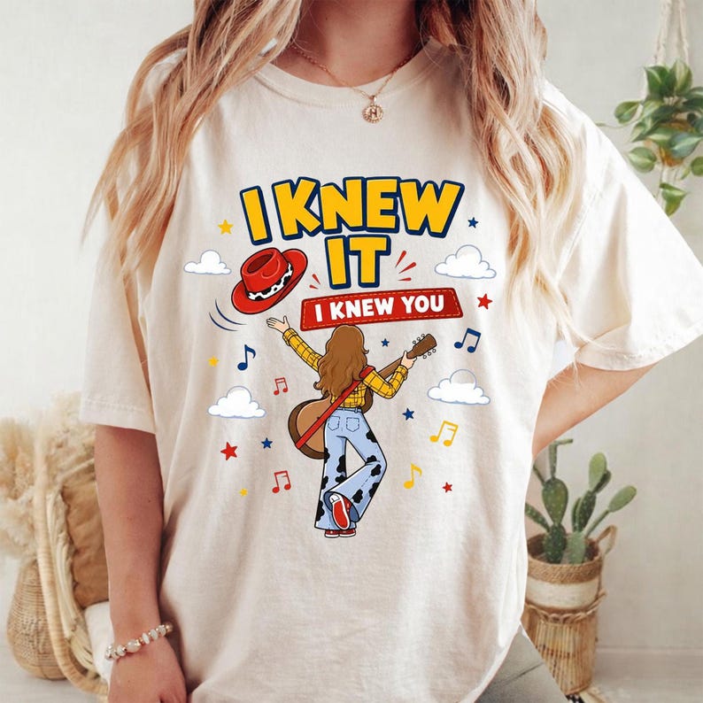 Puede incluir: Una camiseta color crema con un gr&aacute;fico de una persona de pelo largo casta&ntilde;o tocando una guitarra ac&uacute;stica. El dise&ntilde;o incluye un sombrero de vaquero rojo, pantalones con estampado de vaca, notas musicales, estrellas, nubes y el texto I KNEW IT I KNEW YOU.