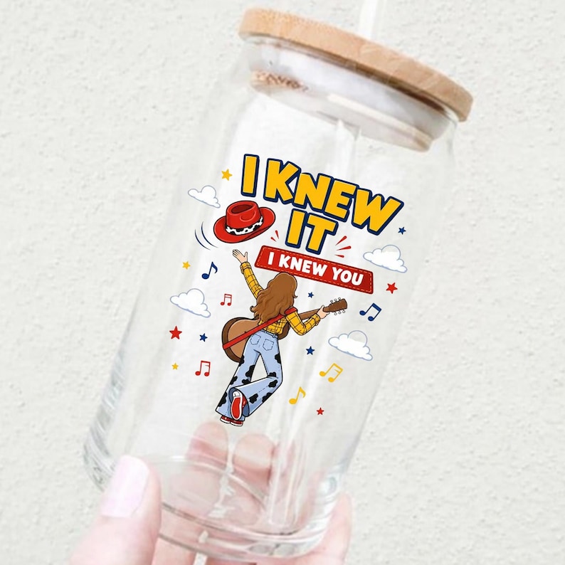 Puede incluir: Un vaso transparente con tapa de bamb&uacute; que presenta un gr&aacute;fico de una persona tocando una guitarra ac&uacute;stica, un sombrero de vaquero rojo y el texto I KNEW IT I KNEW YOU, rodeado de peque&ntilde;as estrellas rojas y amarillas, notas musicales y nubes blancas.
