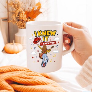 Puede incluir: Una taza de cer&aacute;mica blanca con un gr&aacute;fico colorido de una persona de cabello casta&ntilde;o largo tocando la guitarra, vistiendo una camisa de cuadros amarillos y pantalones con estampado de vaca. El texto dice I KNEW IT y I KNEW YOU con notas.
