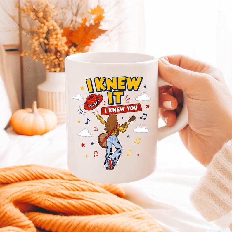 Puede incluir: Una taza de cer&aacute;mica blanca con un gr&aacute;fico colorido de una persona de cabello casta&ntilde;o largo tocando la guitarra, vistiendo una camisa de cuadros amarillos y pantalones con estampado de vaca. El texto dice I KNEW IT y I KNEW YOU con notas.