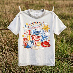 Puede incluir: Una camiseta gris claro de manga corta con un estampado gr&aacute;fico colorido que incluye una bota de vaquero, un sombrero rojo, una estrella de sheriff y el texto: You've Got A Friend In Me, I Knew It, I Knew You, TS5 y To Infinity And Beyond.