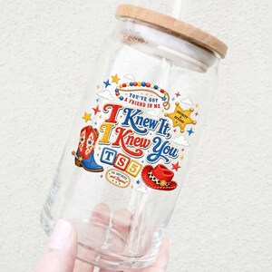 Puede incluir: Un vaso de cristal transparente con tapa de bamb&uacute; y pajita, que presenta un estampado gr&aacute;fico colorido que incluye una bota de vaquero, un sombrero de vaquero, una estrella y el texto "You've Got A Friend In Me", "I Knew It, I Knew You", "TS5" y "To Infinity And Beyond".