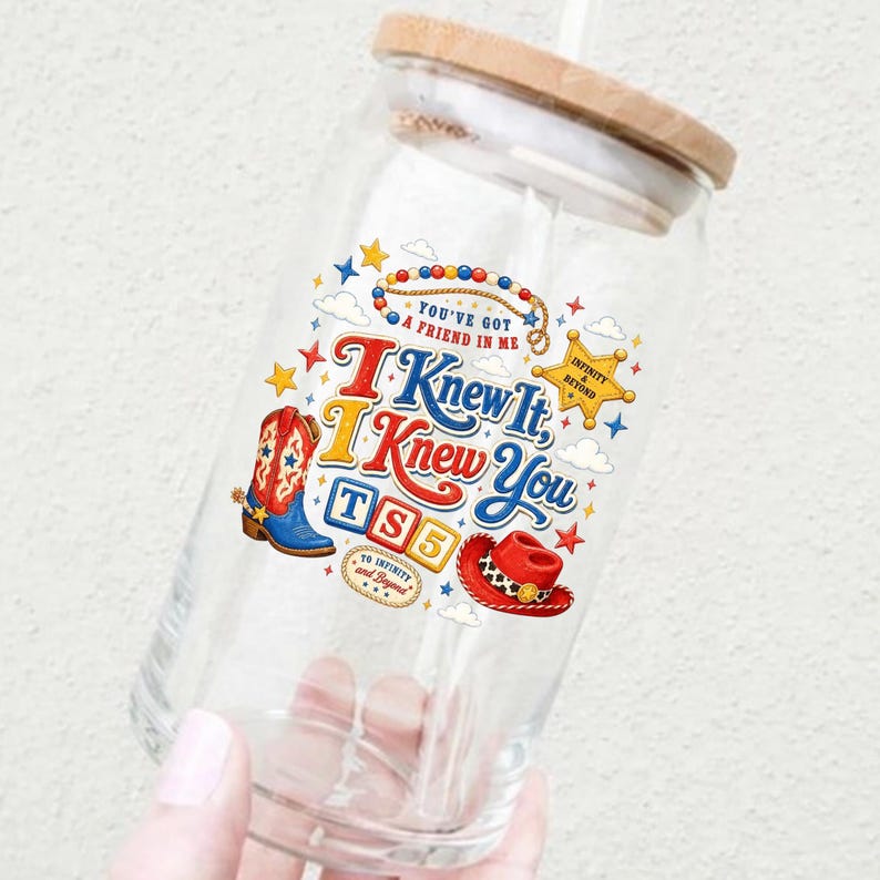 Puede incluir: Un vaso de cristal transparente con tapa de bamb&uacute; y pajita, que presenta un estampado gr&aacute;fico colorido que incluye una bota de vaquero, un sombrero de vaquero, una estrella y el texto "You've Got A Friend In Me", "I Knew It, I Knew You", "TS5" y "To Infinity And Beyond".