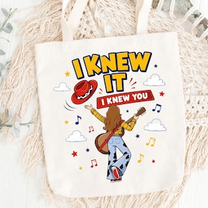 Puede incluir: Una bolsa de tela blanca con un gr&aacute;fico de una persona de pelo largo casta&ntilde;o con camisa de cuadros amarilla y pantalones acampanados con estampado de vaca, sosteniendo una guitarra. El texto dice I KNEW IT y I KNEW YOU, con notas musicales.