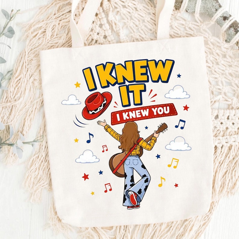Puede incluir: Una bolsa de tela blanca con un gr&aacute;fico de una persona de pelo largo casta&ntilde;o con camisa de cuadros amarilla y pantalones acampanados con estampado de vaca, sosteniendo una guitarra. El texto dice I KNEW IT y I KNEW YOU, con notas musicales.