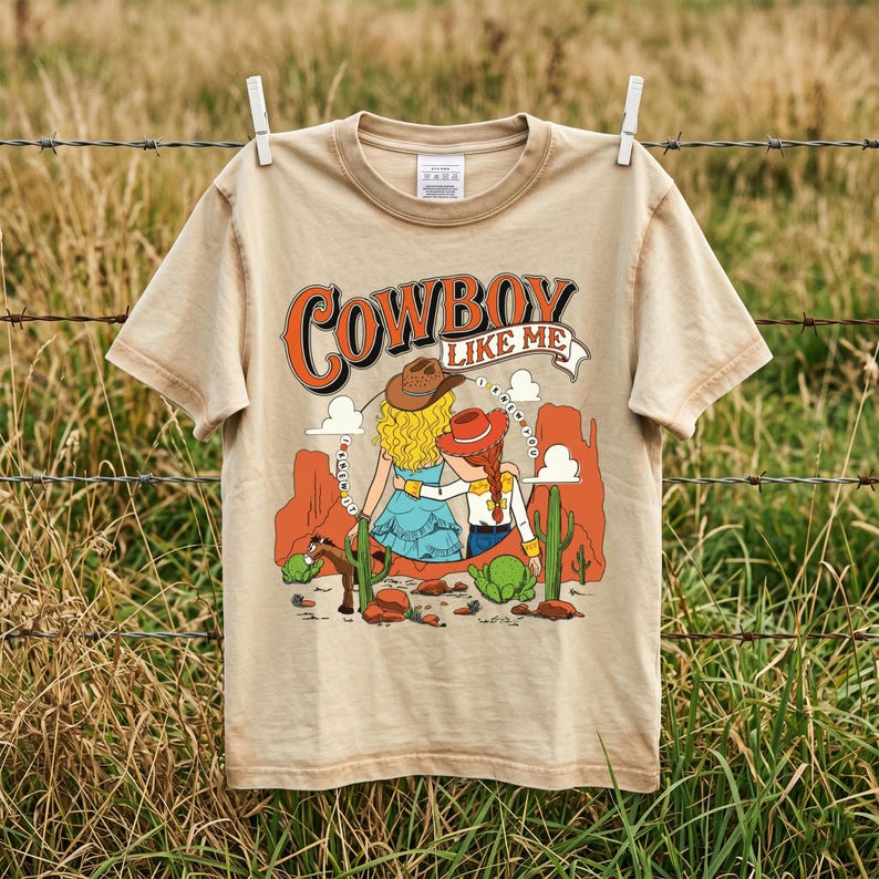 Puede incluir: Una camiseta beige de manga corta con un estampado gr&aacute;fico de dos figuras con sombreros de vaquero y el texto "COWBOY LIKE ME". La ilustraci&oacute;n incluye formaciones rocosas naranjas, cactus verdes y un caballo peque&ntilde;o en un desierto.
