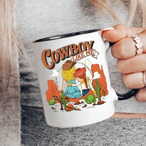 Puede incluir: Una taza de cer&aacute;mica blanca con asa y borde negros, que presenta una ilustraci&oacute;n gr&aacute;fica de dos figuras con sombreros de vaquero en un paisaje des&eacute;rtico con cactus y formaciones rocosas rojas, con el texto COWBOY LIKE ME impreso arriba.