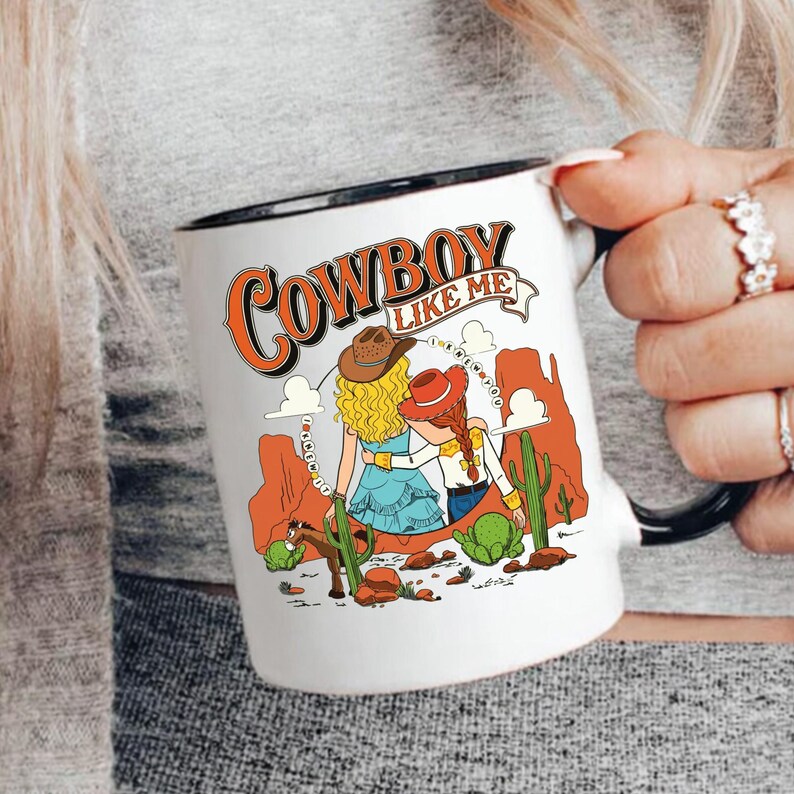 Puede incluir: Una taza de cer&aacute;mica blanca con asa y borde negros, que presenta una ilustraci&oacute;n gr&aacute;fica de dos figuras con sombreros de vaquero en un paisaje des&eacute;rtico con cactus y formaciones rocosas rojas, con el texto COWBOY LIKE ME impreso arriba.