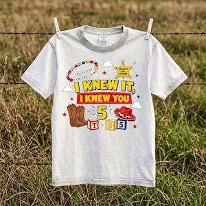 Pu&ograve; includere: Una t-shirt a maniche corte di colore chiaro con una stampa grafica che include il testo I KNEW IT, I KNEW YOU, il numero 5, una stella da sceriffo, un cappello da cowboy, stivali marroni, blocchi con lettere e perline colorate.