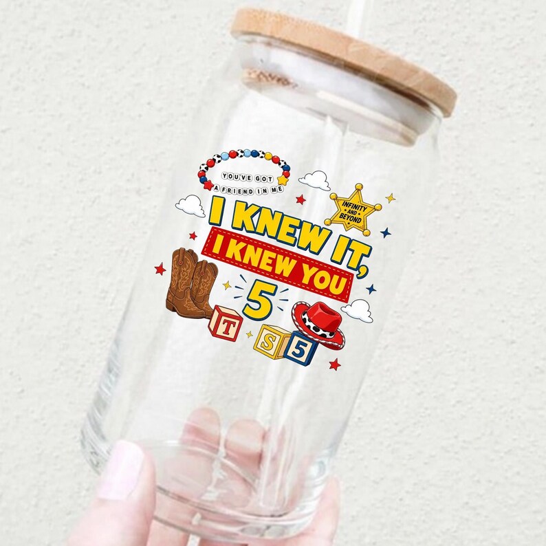 Pu&ograve; includere: Un bicchiere trasparente con coperchio in bamb&ugrave; caratterizzato da una stampa grafica colorata. Il design include stivali da cowboy marroni, un cappello da cowboy rosso, blocchi con le lettere T, S e 5, una stella da sceriffo dorata e il testo I KNEW IT, I KNEW YOU.