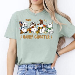 May include: A sage green short-sleeve t-shirt featuring a graphic of four white ghosts dressed in scarves and holding books, a broom, and a golden cup. The text "HARRY GHOSTER" is printed in orange below the ghosts, flanked by two lightning bolt icons.