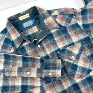 May include: A long-sleeve button-down shirt featuring a plaid pattern in shades of teal, cream, and brown. The shirt has a dark green interior lining, a pointed collar, two chest pockets with flaps, and silver-toned snap buttons on the front.