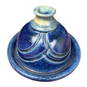 May include: A ceramic butter bell with a deep blue glaze and a lighter beige rim at the top opening. The dome-shaped lid features a series of embossed, curved, wave-like patterns around its exterior, resting securely on a matching circular base.