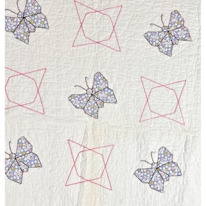 May include: A white quilt with a repeating pattern of embroidered butterflies and geometric shapes. The butterflies are made of a patterned fabric with blue, yellow, and white details. The geometric shapes are outlined in pink thread.