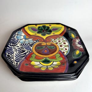 Puede incluir: Una pila de cuatro platos octogonales negros de cerámica. Cada plato presenta un diseño vibrante pintado a mano con patrones florales y geométricos en rojo, amarillo, azul, verde y morado. Los platos tienen un acabado brillante.