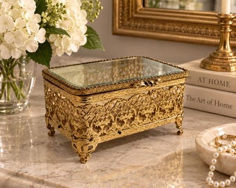 May include: A rectangular jewelry box featuring an intricate gold-tone metal filigree frame with ornate feet and a clear glass lid. The box has a hinged top and a reflective base, showcasing a detailed lattice pattern along all four sides of the container.