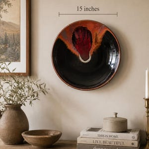 Può includere: Un piatto da parete rotondo in ceramica con un diametro di 15 pollici. Il piatto presenta una smaltatura nera lucida con un motivo strutturato a forma di fiamma rossa e arancione al centro, accentuato da un piccolo dettaglio bianco alla base.
