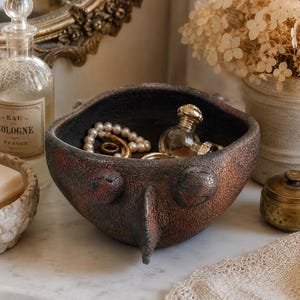 May include: A dark brown, textured ceramic bowl with a rough, crackled finish and a prominent, pointed protrusion on the front. The bowl features two circular, eye-like indentations above the point and contains a string of white pearls and rings.