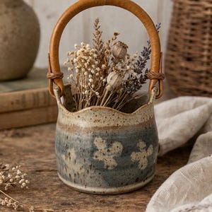May include: A ceramic vase with a brown leather handle, filled with dried flowers. The vase has a blue and tan floral design. The handle is attached to the vase with leather straps. The vase is sitting on a wooden surface.