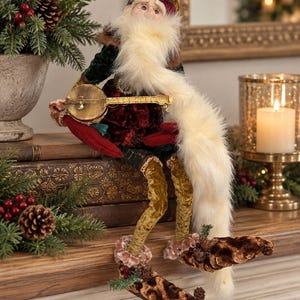 May include: A decorative Santa Claus figurine wearing a dark green and deep red velvet outfit with a long white faux fur beard. The figure holds a small gold-colored banjo and wears pointed brown textured shoes with small pinecone accents.