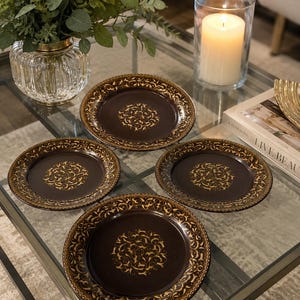 May include: A set of four round plates featuring a dark brown finish with intricate gold-toned filigree patterns centred on the base and along the outer rim. The plates have a smooth, glossy surface and a consistent circular design throughout.