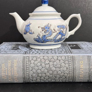 May include: A small, white ceramic teapot with a blue dragon design. The teapot has a curved spout, a loop handle, and a lid with a blue knob. Blue stripes encircle the body. The teapot is resting on a vintage book.