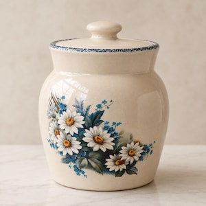 Op de afbeelding: Crèmekleurige keramische pot met deksel, versierd met een bloemenontwerp van witte madeliefjes en blauwe accenten. De pot heeft een blauwe rand rond de rand en een bijpassend deksel.