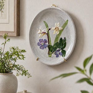 May include: A decorative, oval-shaped ceramic plate with a floral design. The plate features embossed white daffodils, purple flowers, and green leaves. It is mounted on a wall with gold-colored hardware. The plate is a light gray color.