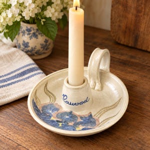 Può includere: Un portacandele in ceramica color crema con manico e un supporto centrale che sostiene una candela affusolata accesa. La base presenta un motivo floreale blu dipinto a mano e la parola "Rauwod" scritta in blu sul supporto centrale.