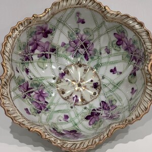 Può includere: Una ciotola antica in porcellana dipinta a mano con bordo smerlato. Presenta un delicato disegno di viole viola e foglie verdi su sfondo bianco, rifinito con bordi dorati. L'interno presenta un motivo floreale centrale.