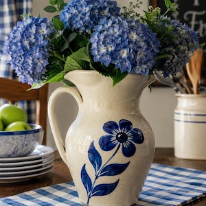 May include: A cream-colored ceramic pitcher with a blue floral design, filled with vibrant blue hydrangea flowers. The pitcher sits on a blue and white checkered tablecloth, creating a rustic, farmhouse aesthetic.
