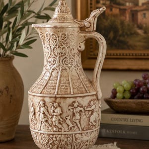 May include: A cream-colored ceramic lidded pitcher featuring intricate relief carvings of figures, foliage, and German text. The vessel has a tall, tapered neck, a curved handle, and a decorative lid with a pointed finial, finished with a brown glaze.