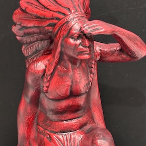 Könnte beinhalten: Eine einfarbige rote Skulptur einer Figur, die einen großen Federschmuck trägt. Die Figur ist in kniender Haltung dargestellt, eine Hand ist an die Stirn gehoben, als würde sie in die Ferne blicken, mit detaillierten Gesichtszügen.