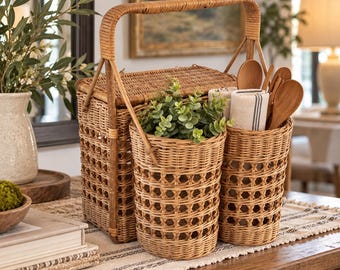 May include: A rectangular woven wicker basket with a tall handle, featuring two attached cylindrical wicker holders. One holder contains a green plant, and the other holds wooden kitchen utensils and a white cloth with black stripes.
