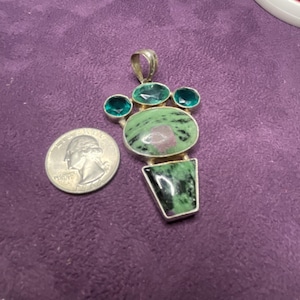 May include: A silver pendant with a large oval green and pink stone, three smaller green stones, and a rectangular green stone. A U.S. quarter is next to the pendant for size comparison. The pendant is on a purple surface.