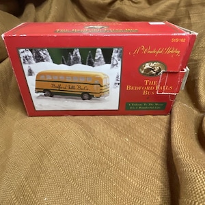 Op de afbeelding: Een rode doos met een model van een gele schoolbus, met het opschrift "Bedford Falls Bus Co.", tegen een winterachtergrond. De doos toont ook de tekst "A Wonderful Holiday" en "The Bedford Falls Bus".