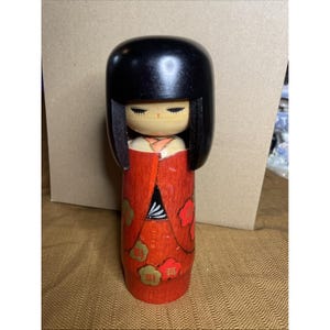 Puede incluir: Muñeca Kokeshi de madera con cabeza negra redondeada y kimono rojo. El kimono está decorado con motivos florales dorados y rojos. La cara de la muñeca tiene rasgos delicados, incluyendo ojos cerrados y boca pequeña. Artesanía tradicional japonesa.