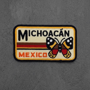 May include: A rectangular embroidered patch with a cream background and a black border. It features the black text MICHOACÁN above horizontal brown, red, and yellow stripes, with the orange text MEXICO below. A butterfly graphic is on the right.