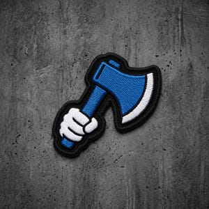 May include: An embroidered patch featuring a cartoon-style white gloved hand gripping the handle of a blue axe with a white blade edge. The design is outlined in thick black thread, creating a bold and distinct graphic element for fabric application.