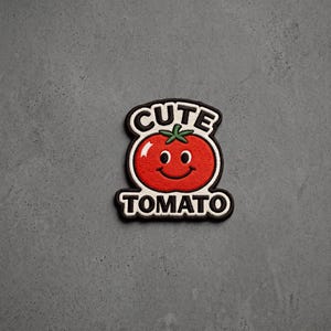 May include: An embroidered patch featuring a bright red tomato with a smiling face, green stem, and white highlights. The text CUTE is arched above the tomato and TOMATO is written below in bold black letters, all outlined in a thick black border.