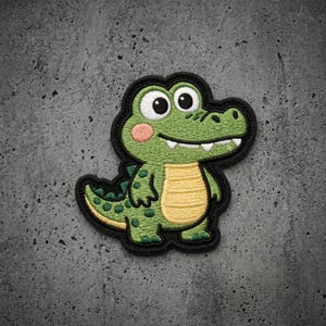May include: An embroidered patch featuring a cartoon crocodile with bright green skin, a yellow belly, and large white eyes. The crocodile has dark green spots on its back and tail, pink cheeks, and a black border outlining the entire shape.
