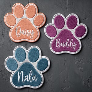 May include: Three embroidered paw print patches with white borders. One patch is peach with the name Daisy, one is purple with the name Buddy, and one is teal with the name Nala. Each patch features white script lettering centered on the paw pad.
