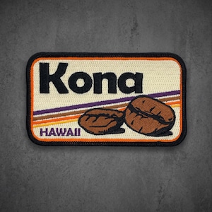 May include: A rectangular embroidered patch with a cream background, featuring the black text Kona and HAWAII. Two brown coffee beans are depicted on the right side, accented by thin, diagonal purple, brown, and orange stripes across the center.