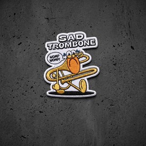 May include: An embroidered patch featuring a cartoon trombone with a sad face, blue teardrops, and the text SAD TROMBONE and WOMP WOMP! The trombone is gold with a white border, and the design is set against a white background with black outlines.