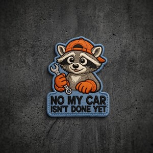 May include: An embroidered patch featuring a cartoon raccoon wearing an orange baseball cap and orange gloves, holding a silver wrench. Below the raccoon, a light blue rectangular banner displays the black text: NO MY CAR ISN'T DONE YET.