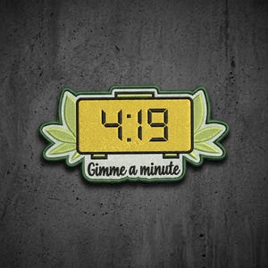 May include: An embroidered patch featuring a yellow digital alarm clock displaying the time 4:19. The clock is flanked by light green leaf shapes on both sides. Below the clock, the text Gimme a minute is stitched in black cursive font.