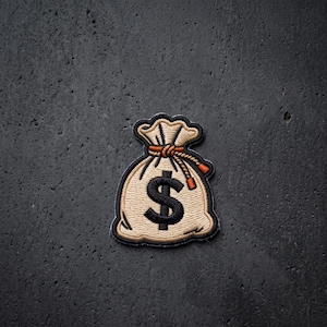 May include: An embroidered patch shaped like a money bag in shades of beige and tan, featuring a prominent black dollar sign symbol in the center and a tied orange drawstring at the top, outlined with a thick black border for clear definition.